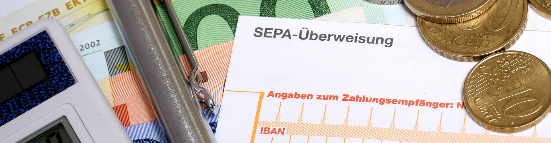 Auf mehreren Geldscheinen liegen Taschenrechner, Kugelschreiber, einige Euromünzen und ein Erlagschein. Zu erkennen ist die Aufschrift "SEPA-Überweisung".