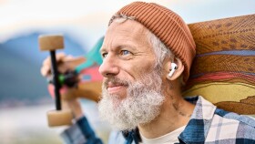 Reiferer, b&auml;rtiger Mann mit M&uuml;tze und Skateboard l&auml;chelt zufrieden