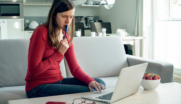 Frau sitzt auf Sofa, arbeitet konzentriert am Laptop, h&auml;lt Stift in der Hand, helle moderne Wohnung.