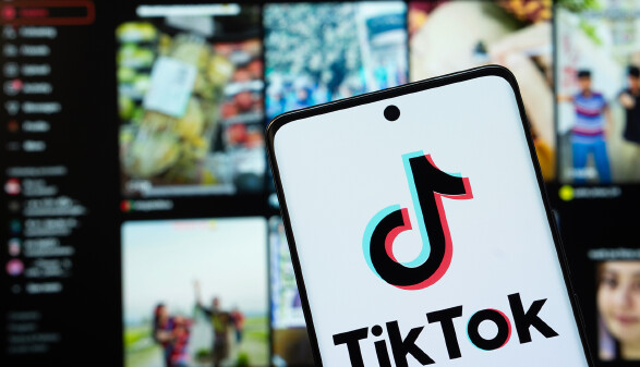 Das TikTok-Logo wird auf einem Smartphone-Bildschirm angezeigt, im Hintergrund ist die TikTok-Website auf einem Computerbildschirm zu sehen