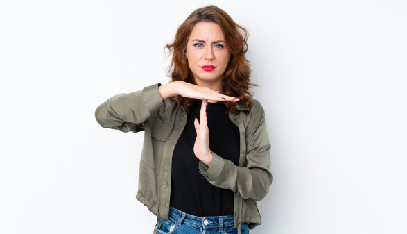 Frau mit welligem braunen Haar und ernstem Blick steht vor einer weißen Wand und macht mit ihren Händen das Zeichen für Auszeit.