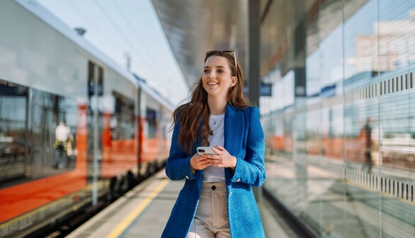 Junge Frau mit Smartphone am Bahnhof auf dem Weg zur Arbeit. Symbolbild f&uuml;r Pendeln und die Nutzung &ouml;ffentlicher Verkehrsmittel.