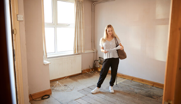 Junge Frau steht in einem renovierungsbedürftigen, leeren Zimmer und schaut Papiere durch