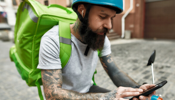 Essenslieferant mit Helm und gro&szlig;em gr&uuml;nen Thermo-Rucksack sucht auf seinem Handy, sitzt dabei auf einem (E-)Moped