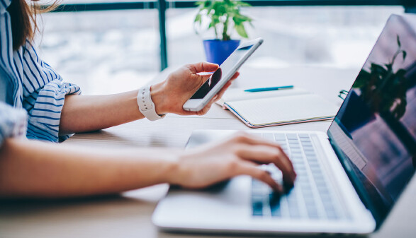 Frau sitzt mit Smartphone in der Hand vor einem Laptop